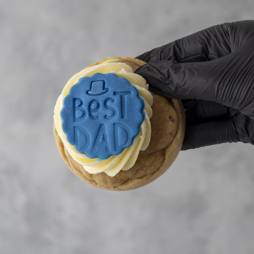 Hand holding a luxury cookie with 'Best Dad' icing against a gray background