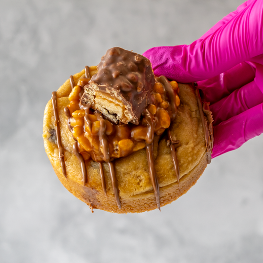 Lion Bar Stuffed Deluxe Cookie with chocolate drizzle held by a gloved hand on a gray background