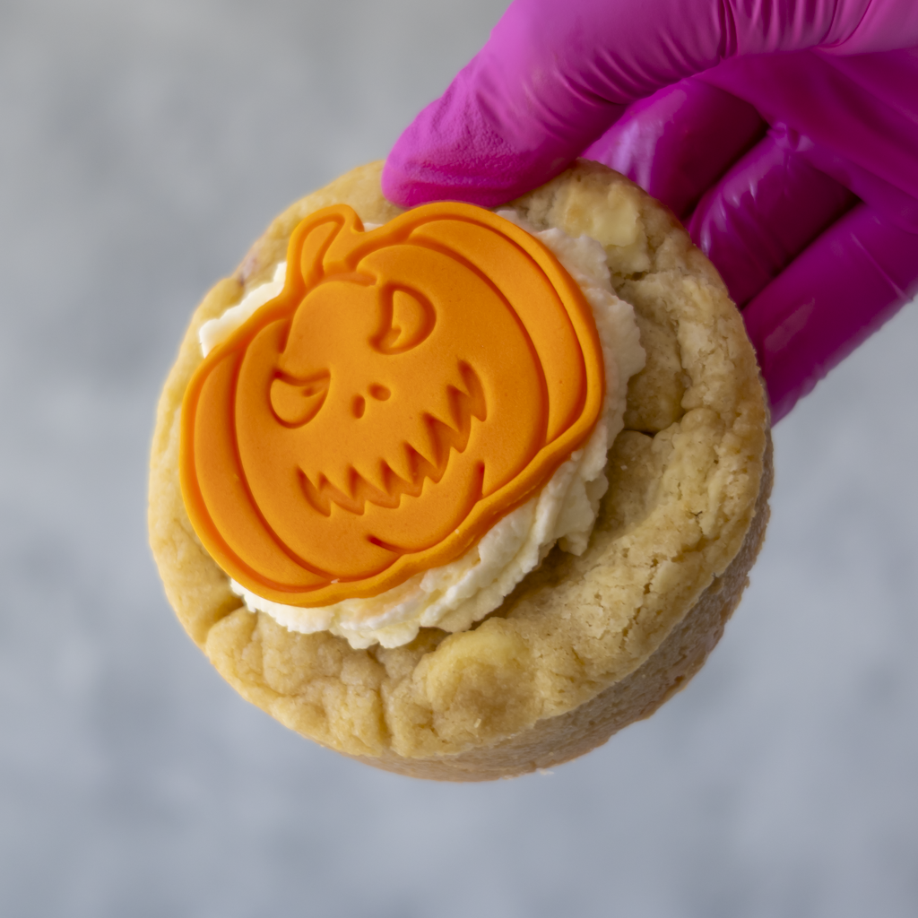 A close-up of a hand wearing a bright magenta glove holding a Little Pumpkin Deluxe Cookie with white frosting and an orange pumpkin-shaped decoration on top, against a soft grey background.
