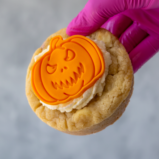 A close-up of a hand wearing a bright magenta glove holding a Little Pumpkin Deluxe Cookie with white frosting and an orange pumpkin-shaped decoration on top, against a soft grey background.