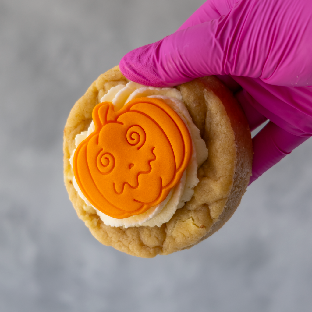 A hand wearing a bright pink glove holds a DoughGirl Little Pumpkin Deluxe Cookie topped with white frosting and an orange pumpkin-shaped decoration, against a soft grey background.