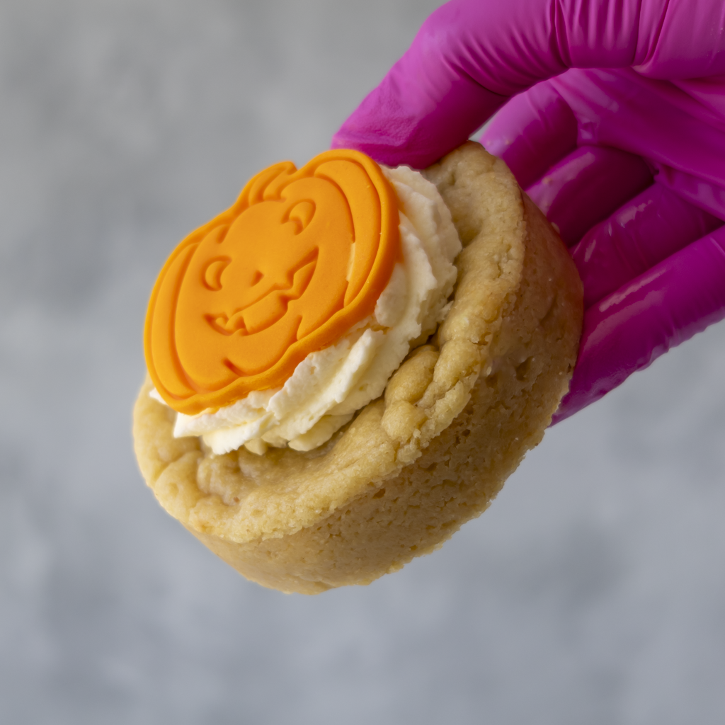 A hand wearing a bright magenta glove holds a small pumpkin-shaped orange decoration on top of white frosting on a round cookie, against a soft gray background.