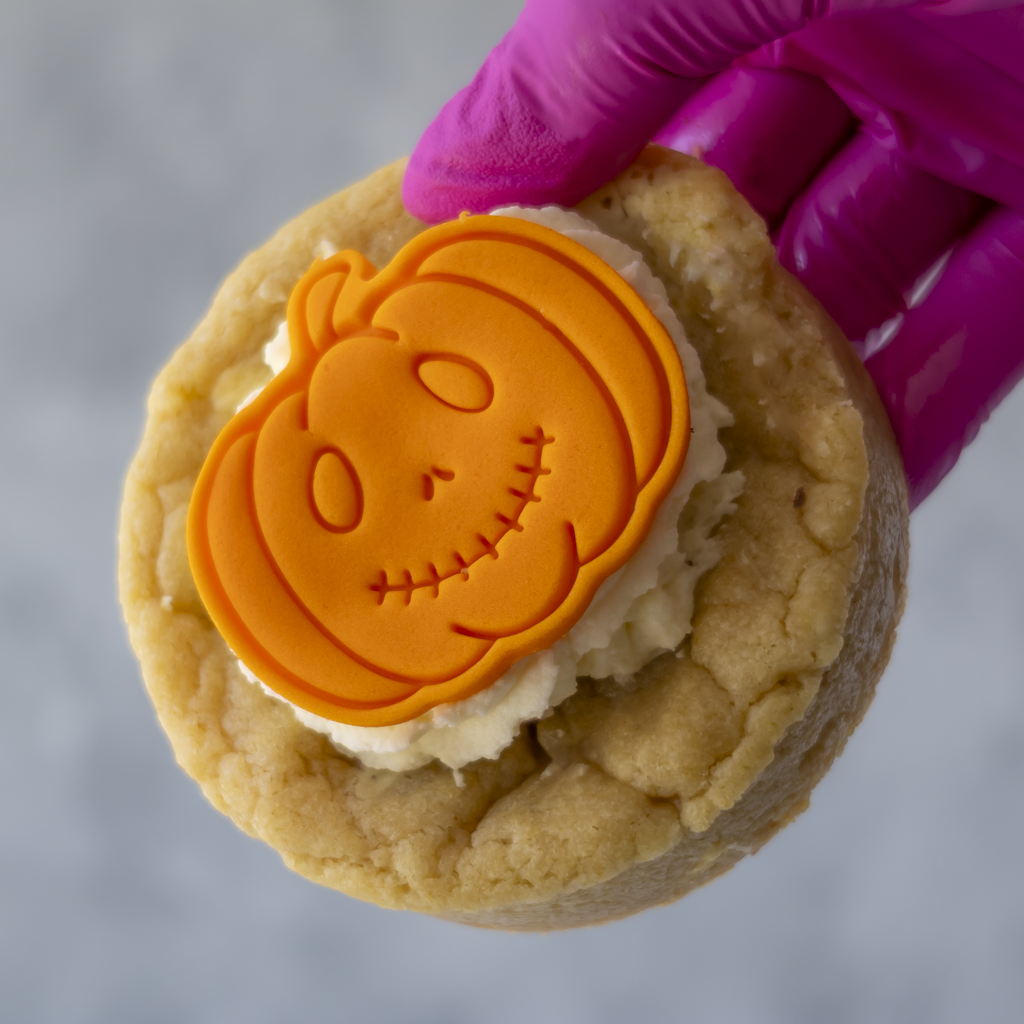A close-up of a hand wearing a bright pink glove holding a round baked cookie topped with a molded orange pumpkin decoration over white frosting, against a soft gray background.