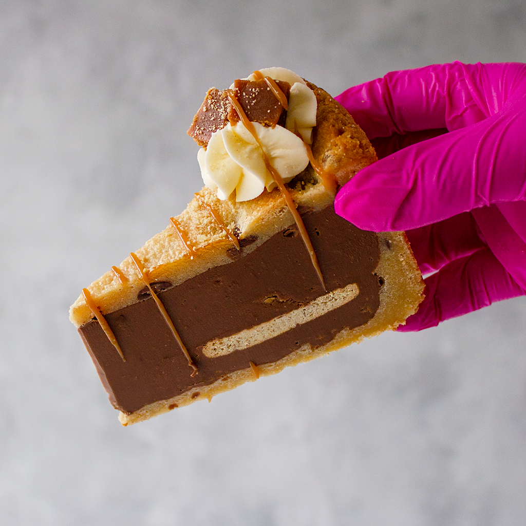 Slice of layered dessert with chocolate and caramel, held by a hand in a pink glove on a neutral background.