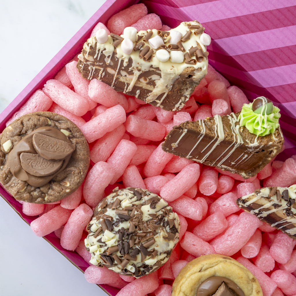 A close-up overhead view of assorted chocolate treats arranged in a pink gift box. The treats include chocolate-covered cookies, a slice of chocolate dessert with white drizzle and mini marshmallows, a slice of chocolate cake with green frosting, and other chocolate pieces.