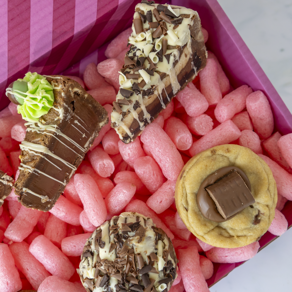 A close-up overhead view of assorted chocolate treats arranged in a pink gift box. The treats include two chocolate cake slices with drizzle and sprinkles, a small green-topped chocolate piece, a chocolate cookie with a chocolate  piece, and a round chocolate-covered cookie topped with chocolate shavings and a chocolate square.