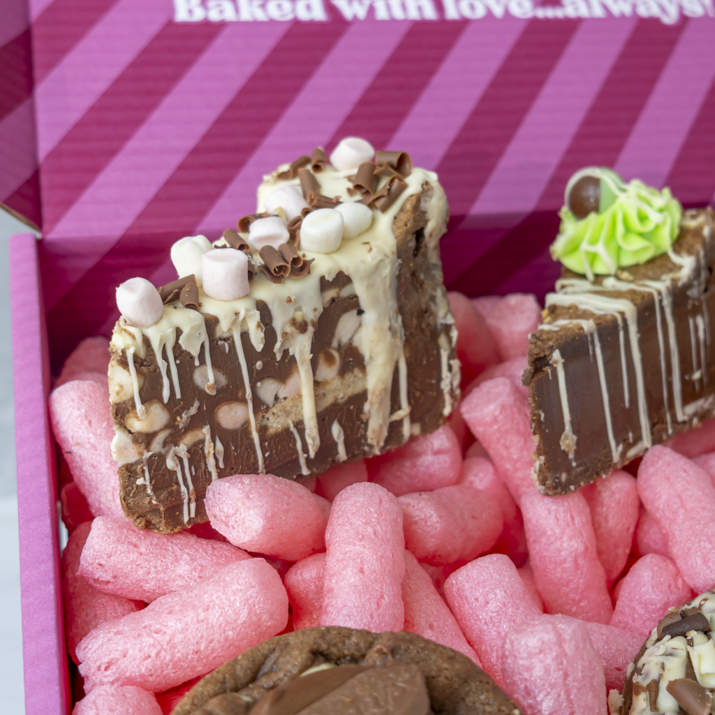 Chocolate chip cookie bars topped with white chocolate drizzle, marshmallows, and chocolate shavings, presented in a pink gift box filled with pink packing peanuts.