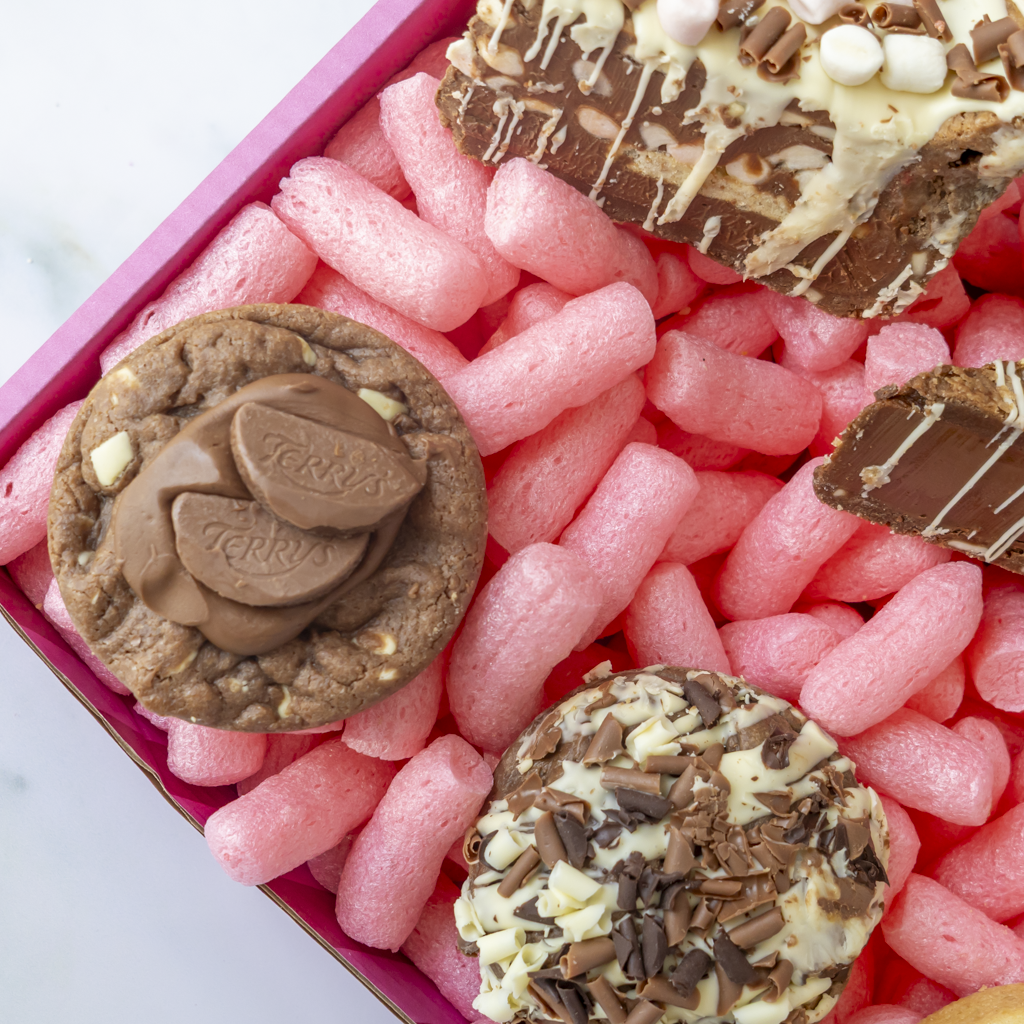 A close-up overhead view of assorted chocolate treats arranged in a pink gift box with pink packing peanuts. Visible items include a round chocolate cookie topped with Terry chocolate pieces, a round chocolate chip cookie with chocolate and white chocolate drizzle, and a chocolate-covered cookie topped with chocolate sprinkles.