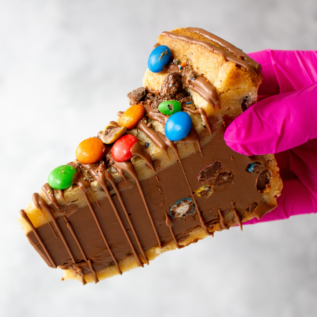 Chocolate-covered cookie pie slidewith colorful M&M candies held by a pink-gloved hand on a white background