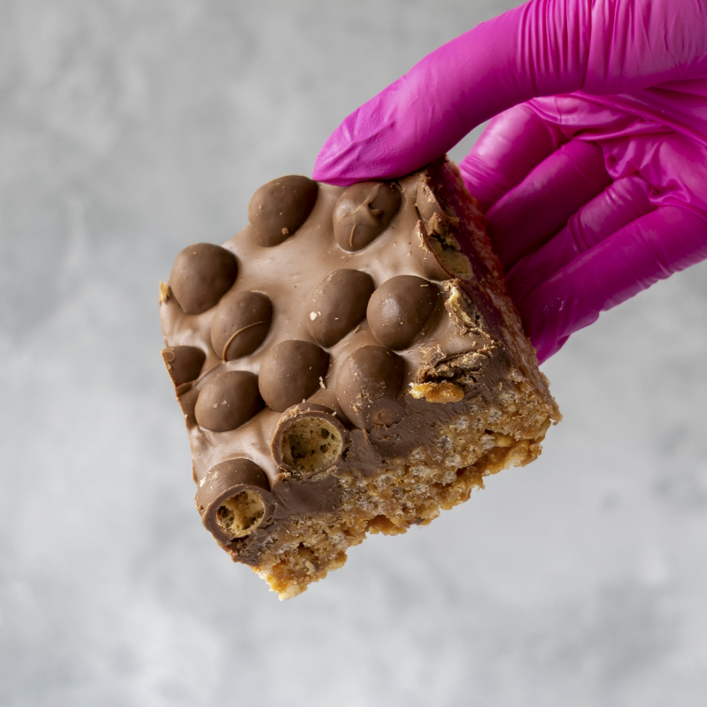 Hand wearing a pink glove holding a chocolate-covered Malteser Crispie Slab snack bar against a neutral background