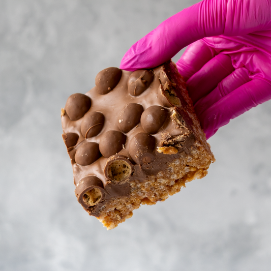 Hand wearing a pink glove holding a chocolate-covered Malteser Crispie Slab snack bar against a neutral background