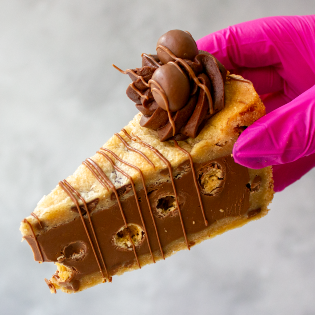 Slice of DoughGirl Malteser Cookie Pie with chocolate drizzle and Malteser pieces held by a hand in a pink glove on a light background