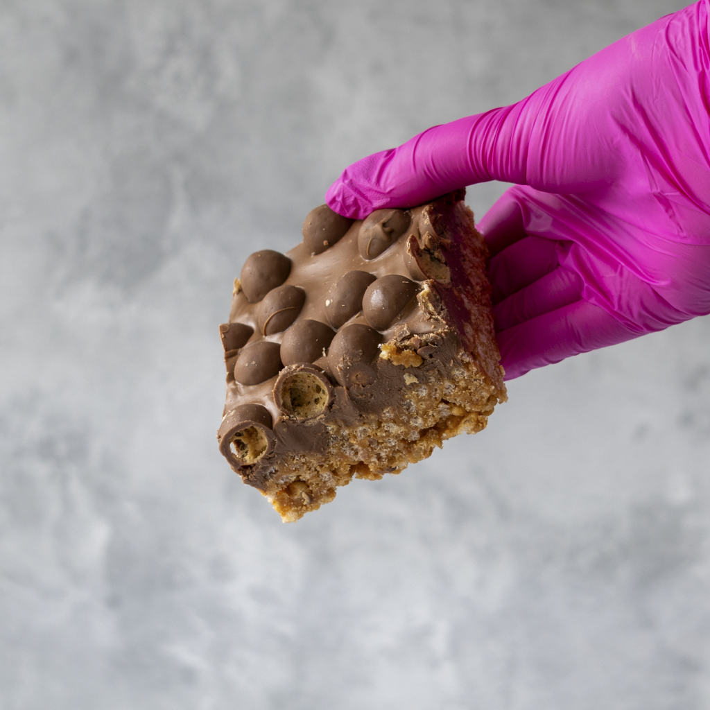 Hand wearing a pink glove holding a Malteser chocolate-covered rice crispy treat against a gray background