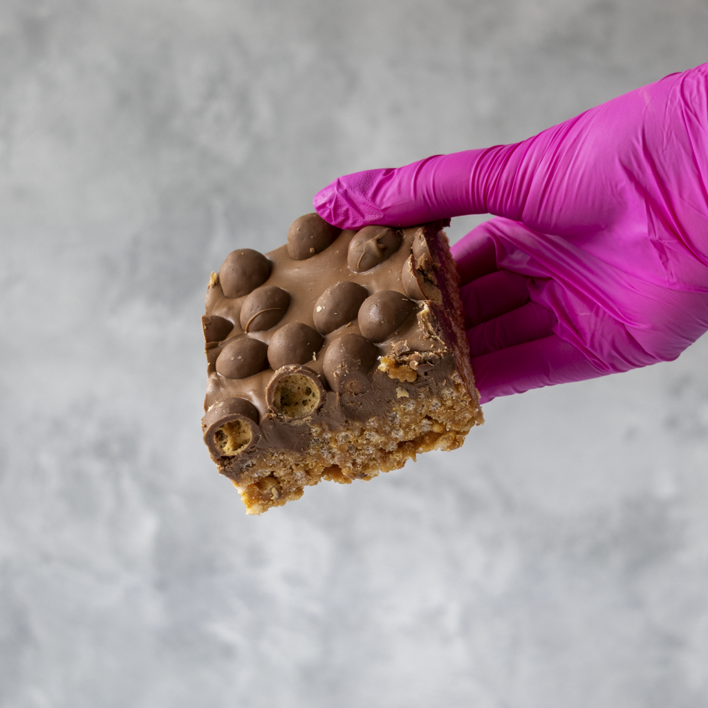 Hand wearing a pink glove holding a Malteser Crispie Slab against a gray background