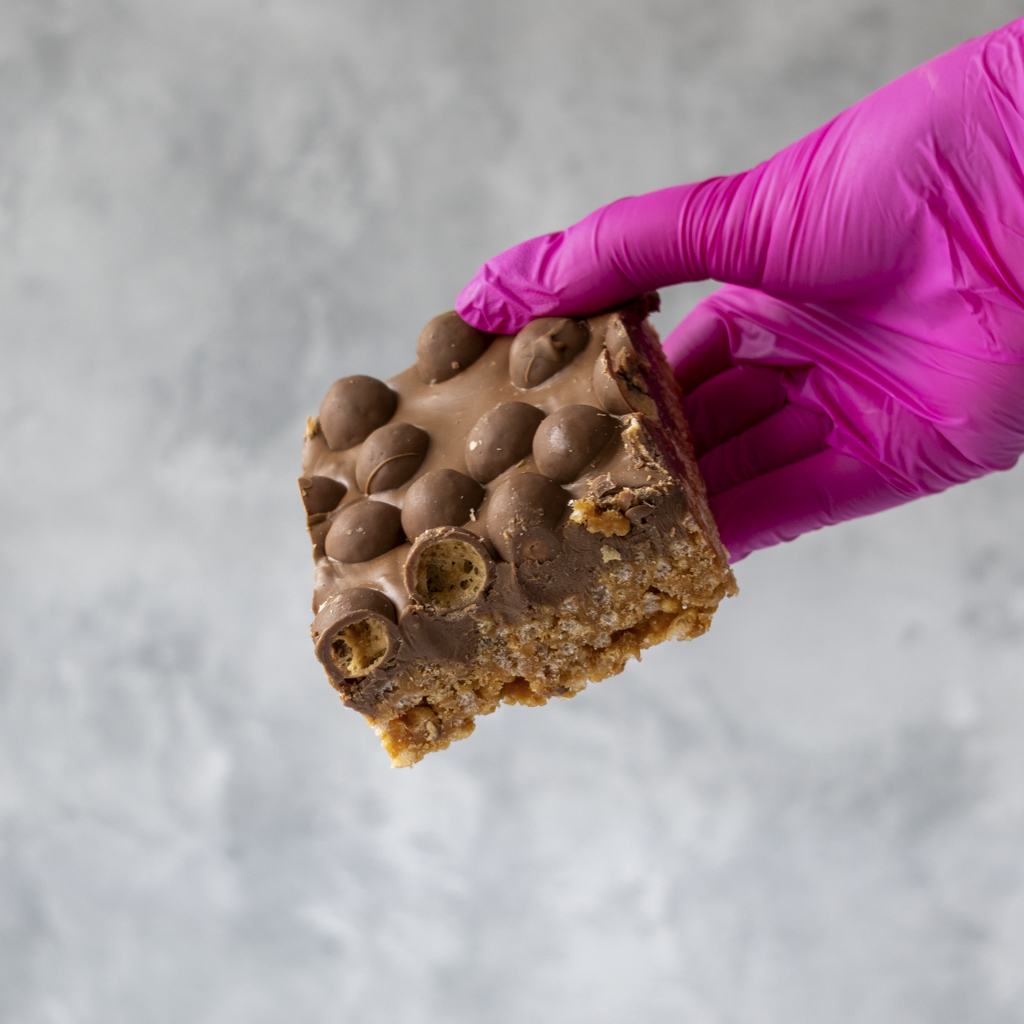 Hand wearing a pink glove holding a Malteser chocolate-covered snack bar against a gray background