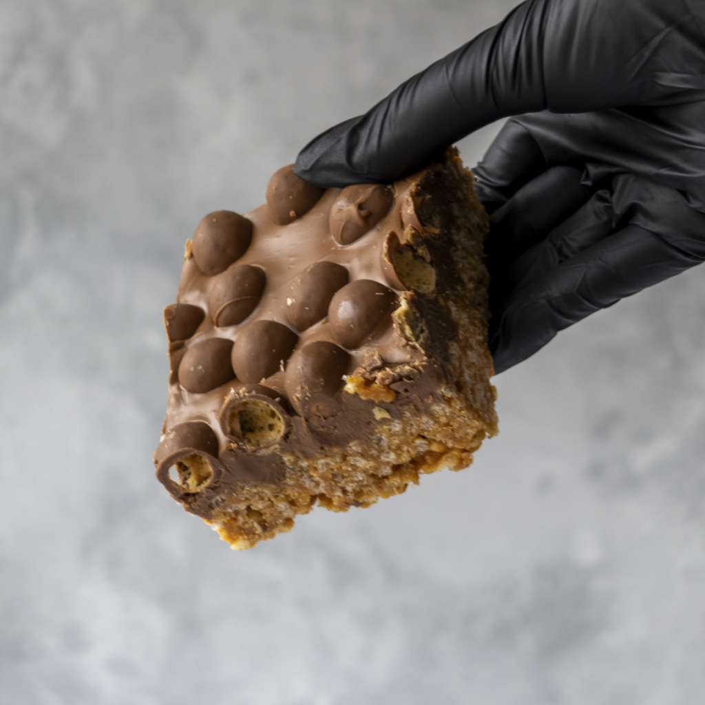 Hand wearing a black glove holding a Malteser Crispie snack bar against a gray background