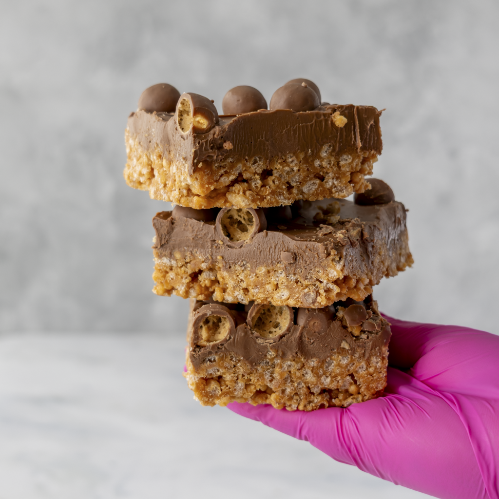 Gloved hand presenting Malteser chocolate crispie slab topped with crispy Malteser balls