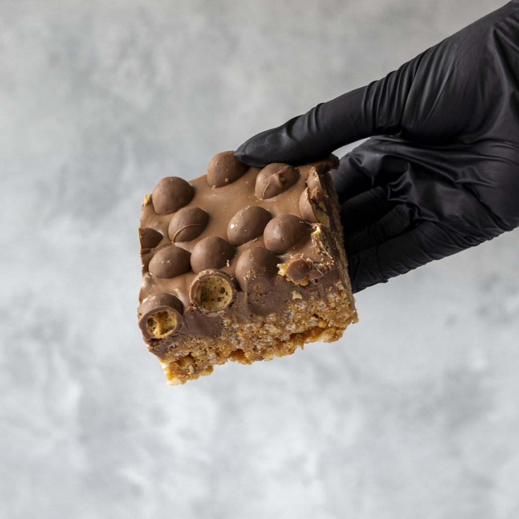 Hand wearing a black glove holding a Malteser Crispie dessert bar against a gray background