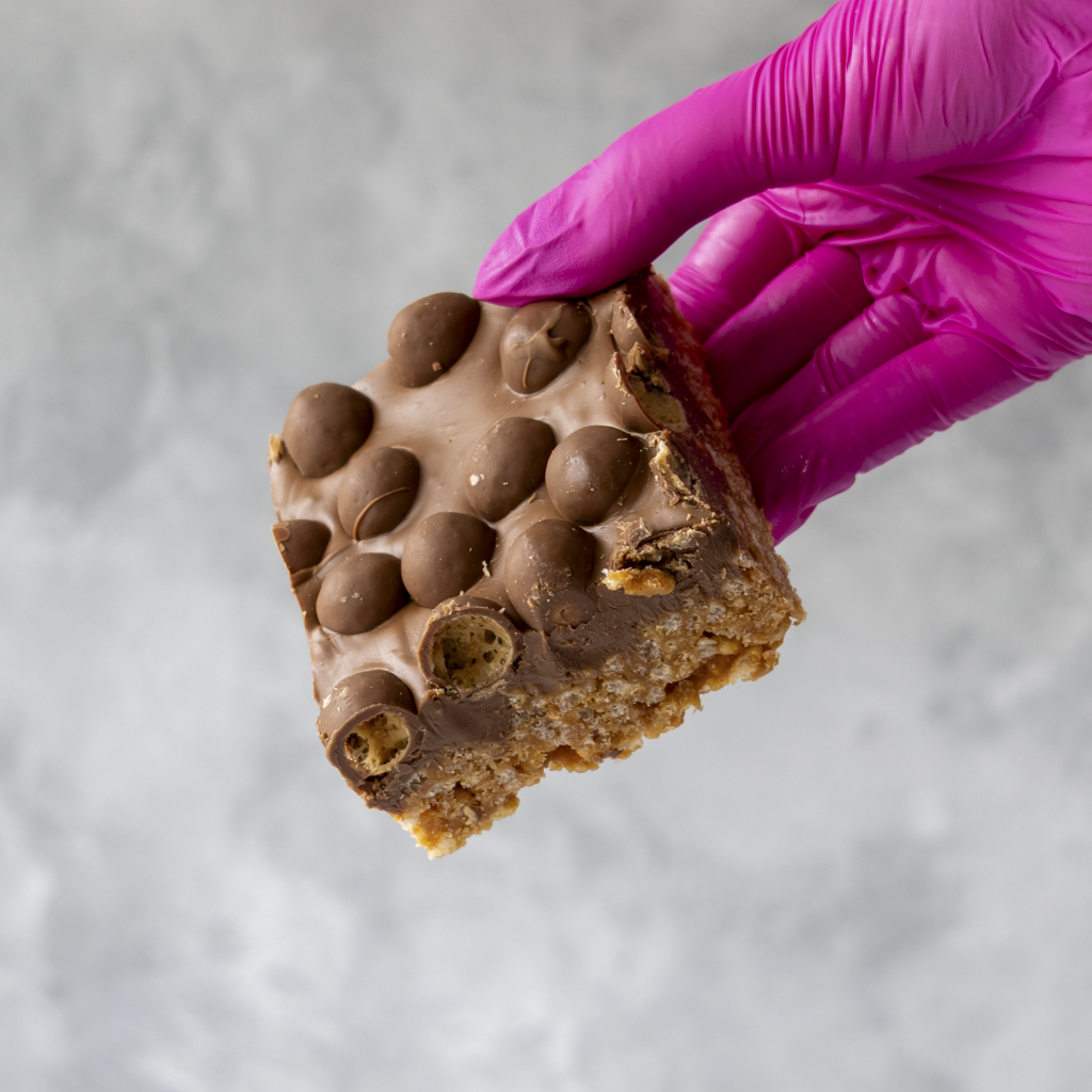 Hand wearing a pink glove holding a Malteser chocolate chip cookie bar against a gray background
