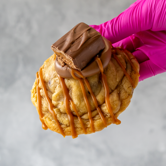 Mars Deluxe Cookie with caramel and chocolate drizzle held by a gloved hand against a neutral background