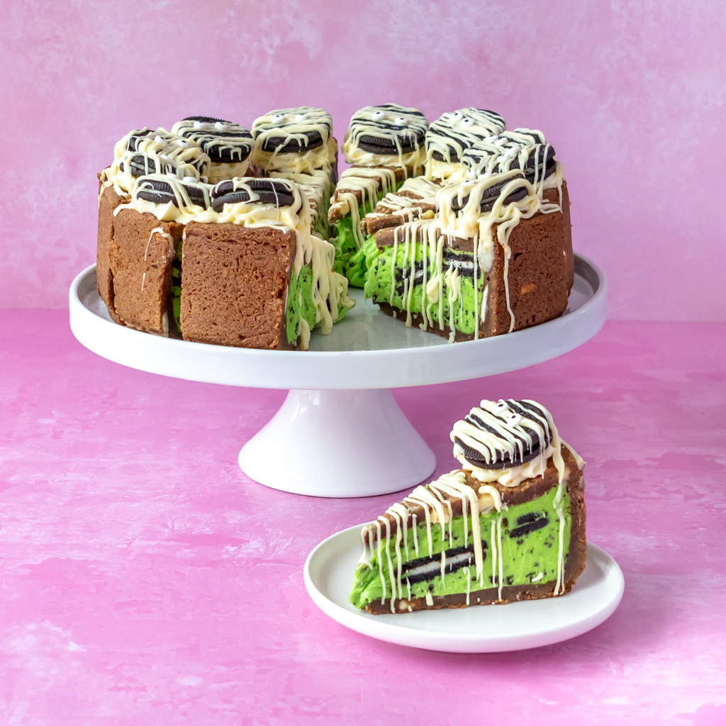 A slice of Midnight Monster Cookies n Cream Pie with chocolate cookie pieces and white drizzle sits on a small white plate in front of a larger matching cake on a white cake stand, set against a pink background.