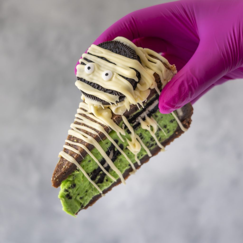A hand wearing a bright pink glove holds a decorated slice of cookie pie shaped like a mummy. The slice has a brown base, green frosting, white chocolate drizzle, and an Oreo cookie on top with candy eyes.