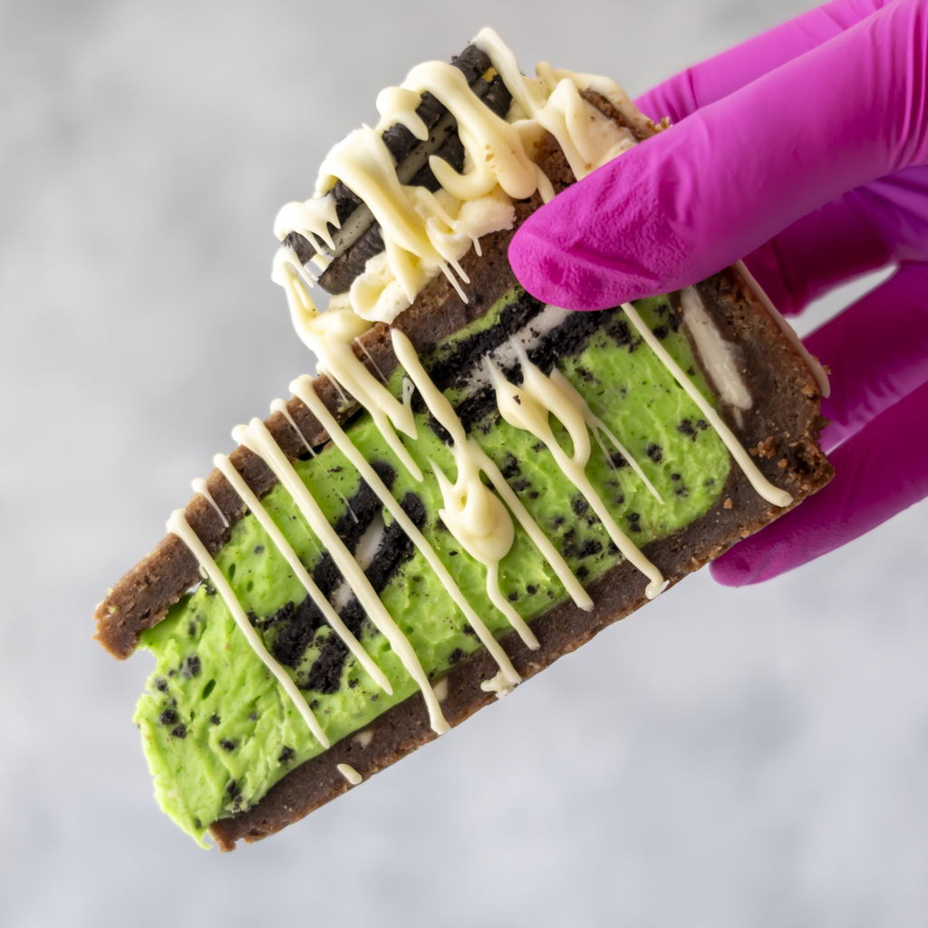 A close-up of a hand wearing a bright pink glove holding a layered dessert bar. The dessert has a brown base, a bright green chocolate with dark speckles, dark cookie pieces, and white chocolate drizzle on top, against a light grey background.