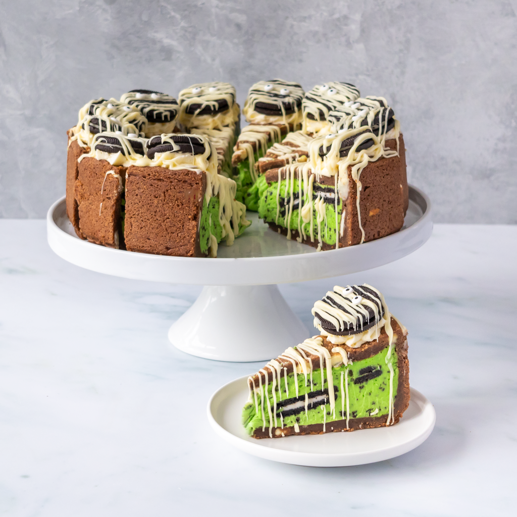 A slice of layered cake on a small white plate in front of a larger cake on a white pedestal stand. The cookie pie has a brown crumb base, bright green cream filling, and white chocolate drizzle with dark cookie pieces on top, set against a light grey background.