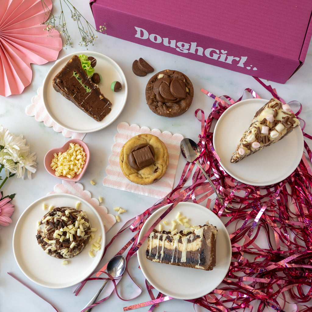 A styled flat-lay of chocolate-dedicated desserts on white plates, with a pink DoughGirl box at the top and pink ribbon scattered around on a light surface.