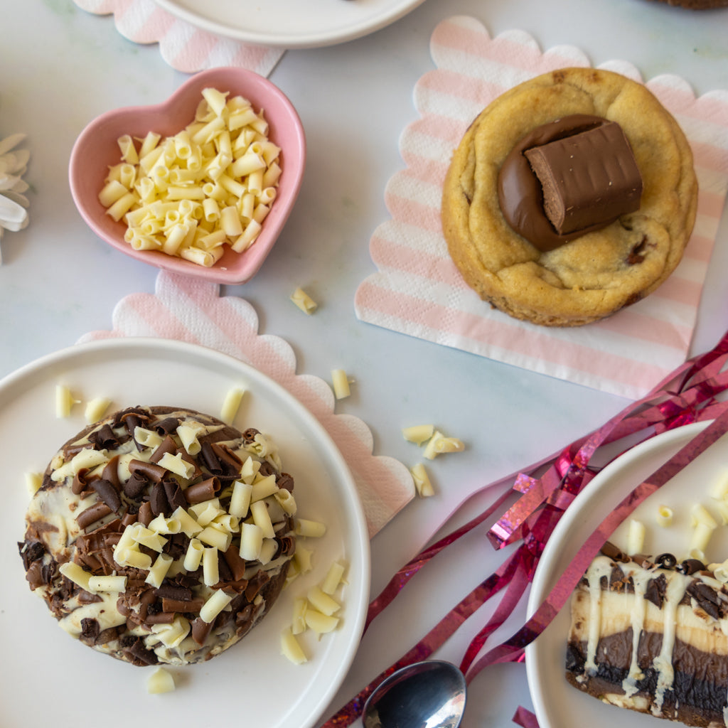A styled flat-lay of chocolate desserts on white plates with pink and white scalloped napkins. One cookie is topped with a milk chocolate bar, and another is covered with dark and white chocolate curls.