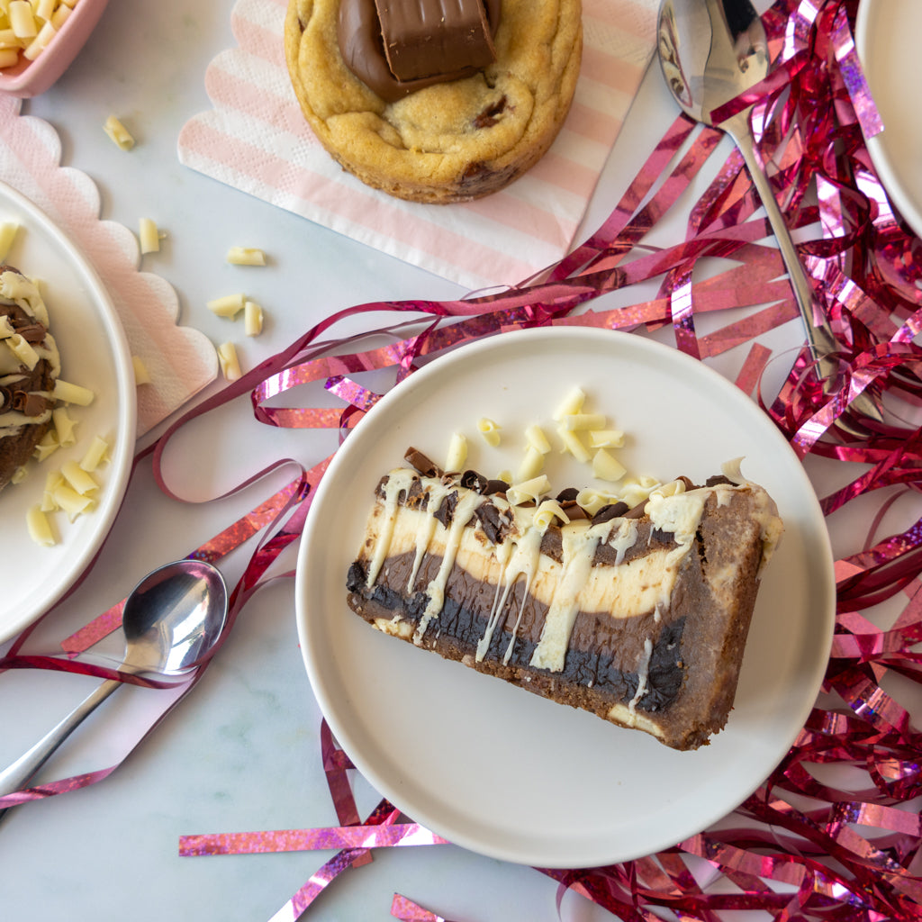 A slice of triple layered chocolate and cream dessert on a white plate, surrounded by pink tinsel and dessert props on a light marble surface.