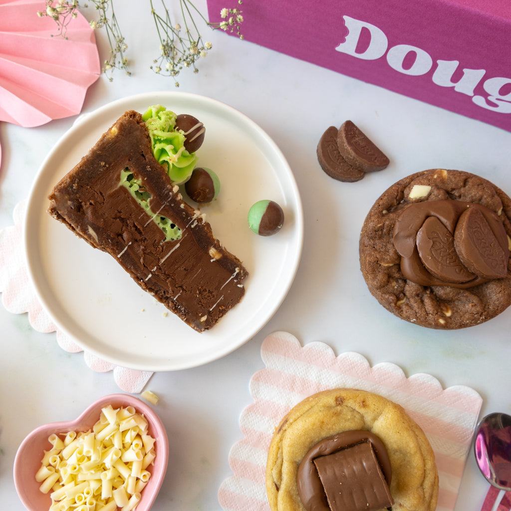 A styled flat-lay of chocolate desserts on a white surface, including a slice of chocolate cake on a white plate, chocolate cookie cups topped with chocolate cookies, chocolate sprinkles, and a pink box with the partial brand name 'DoughGirl'.