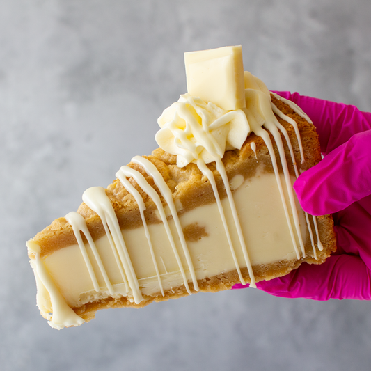 Slice of DoughGirl Milkybar Cookie Pie with white chocolate drizzle held by a hand in a pink glove against a gray background