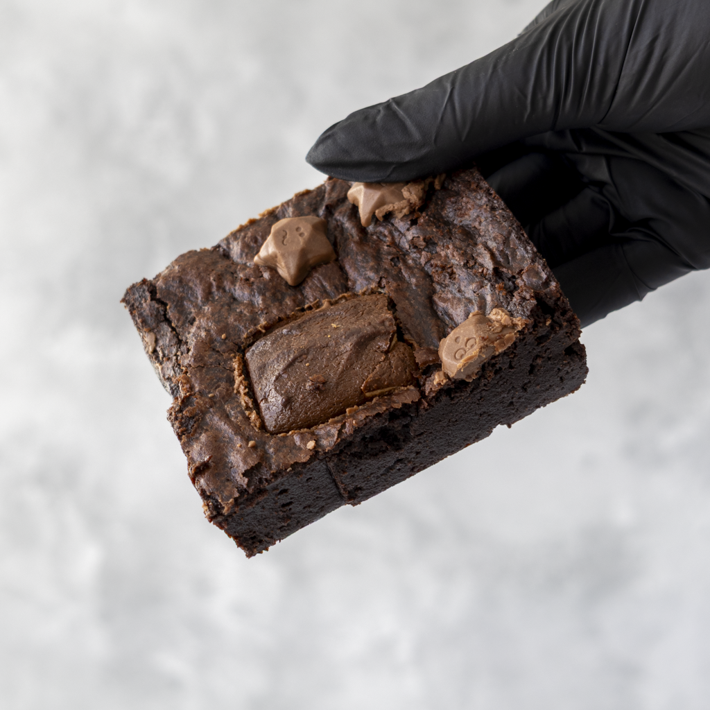 Chocolate brownie with chocolate chips held by a black gloved hand on a light gray background