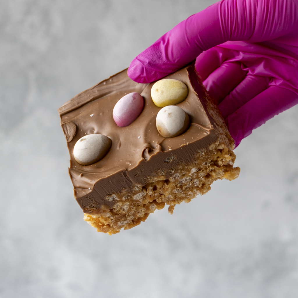 Chocolate-covered treat Mini Egg Crispie Slab with Easter eggs held by a gloved hand on a neutral background