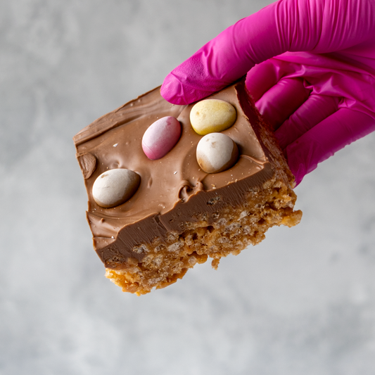 Chocolate-covered treat Mini Egg Crispie Slab with Easter eggs held by a gloved hand on a neutral background