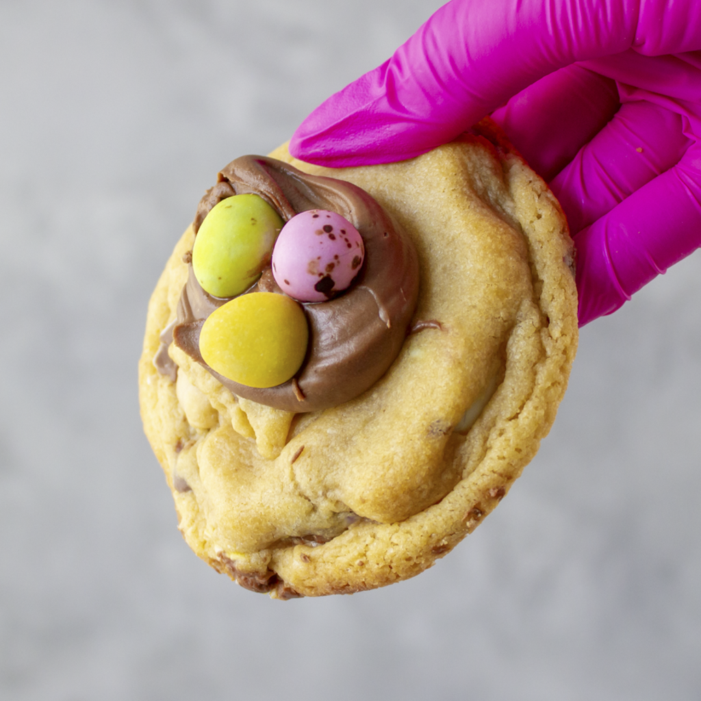 Mini Egg Deluxe Cookie with chocolate frosting and colorful candies held by a gloved hand on a gray background