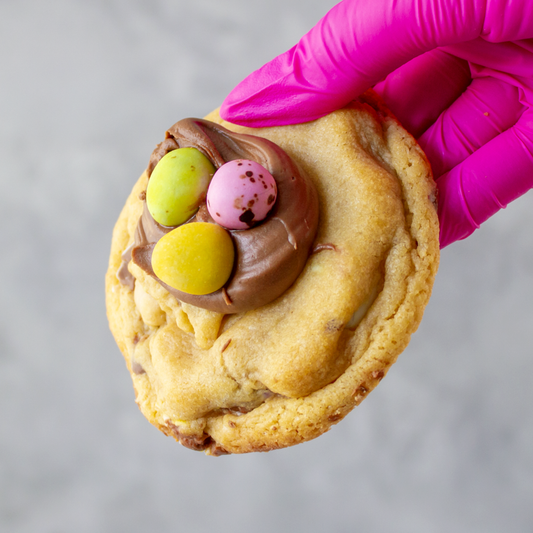 Mini Egg Deluxe Cookie with chocolate frosting and colorful candies held by a gloved hand on a gray background