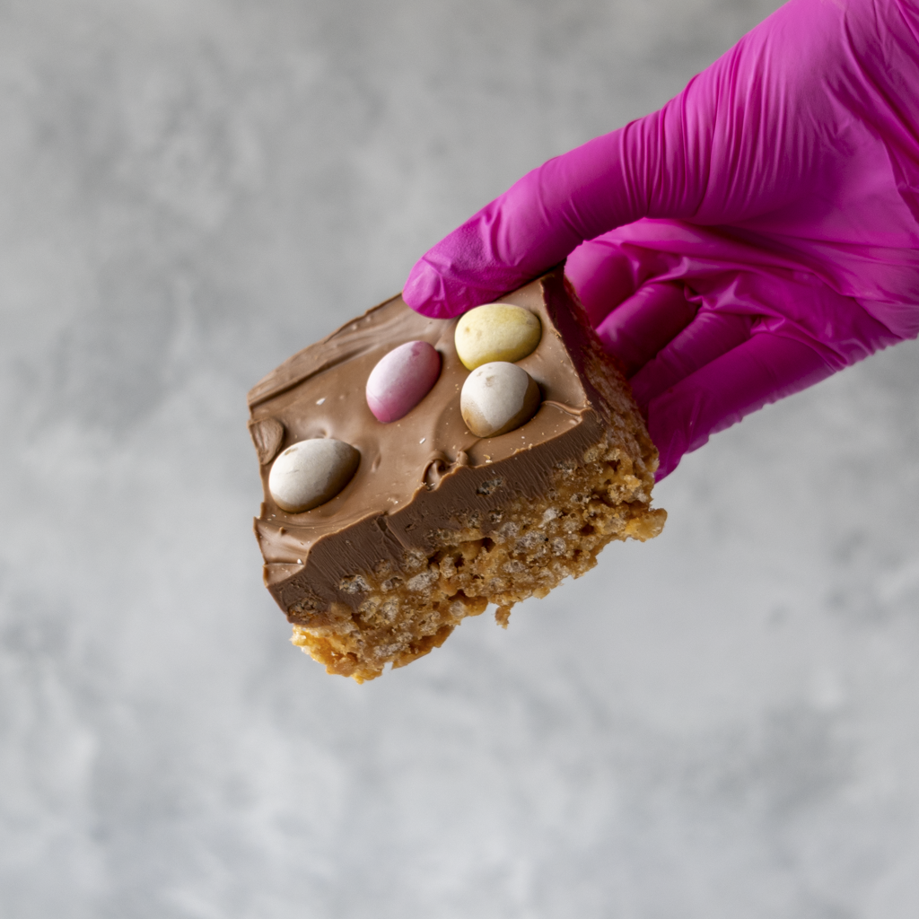 Hand wearing a purple glove holding a piece of Mini Egg Crispie Slab
with Easter eggs on a light gray background