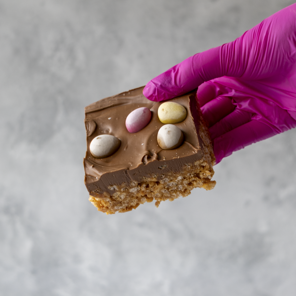 Hand wearing a purple glove holding a Mini Egg Crispie Slab with Easter eggs on a light gray background