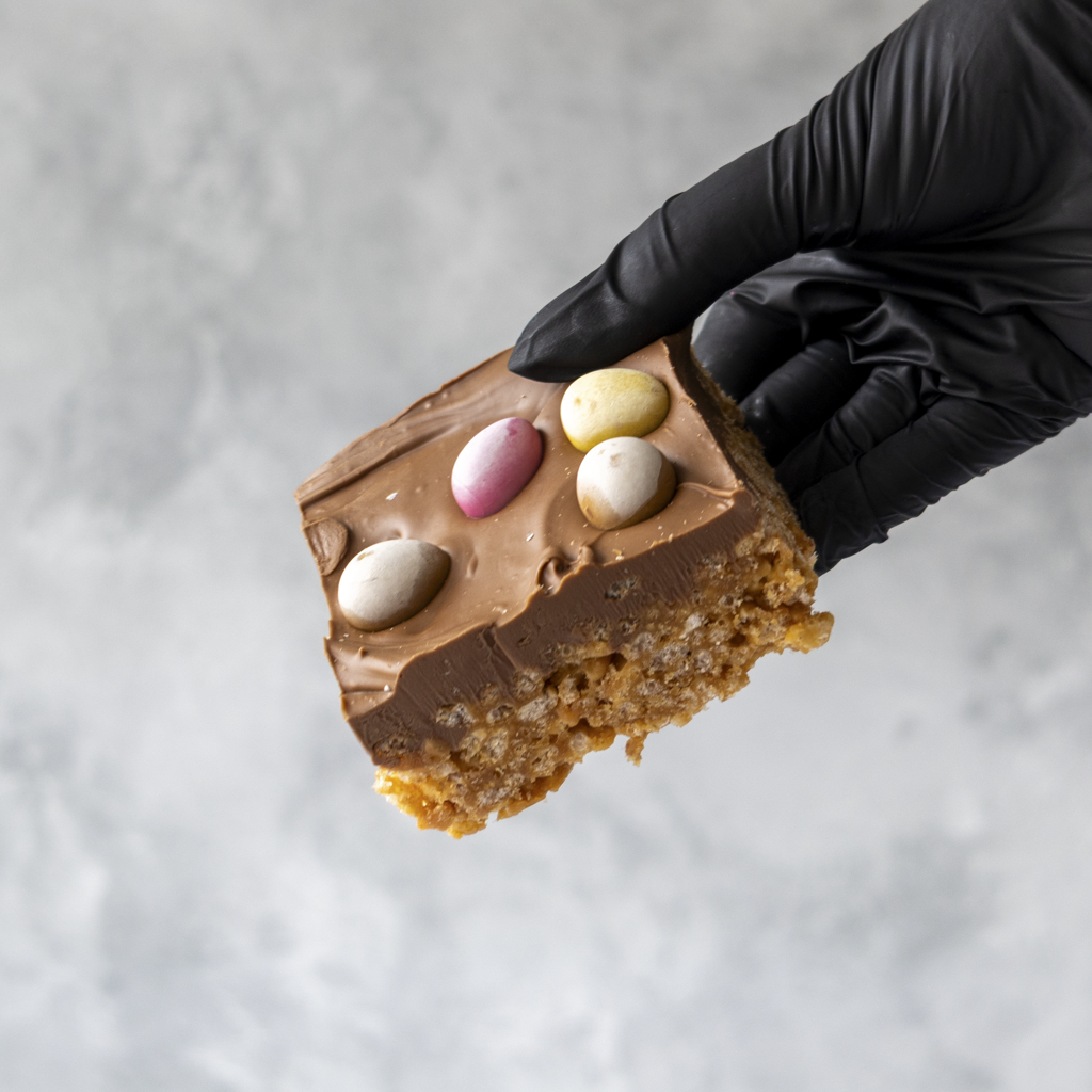 Hand wearing a black glove holding a piece of Crispie Slab with Easter Mini Eggs on a light gray background