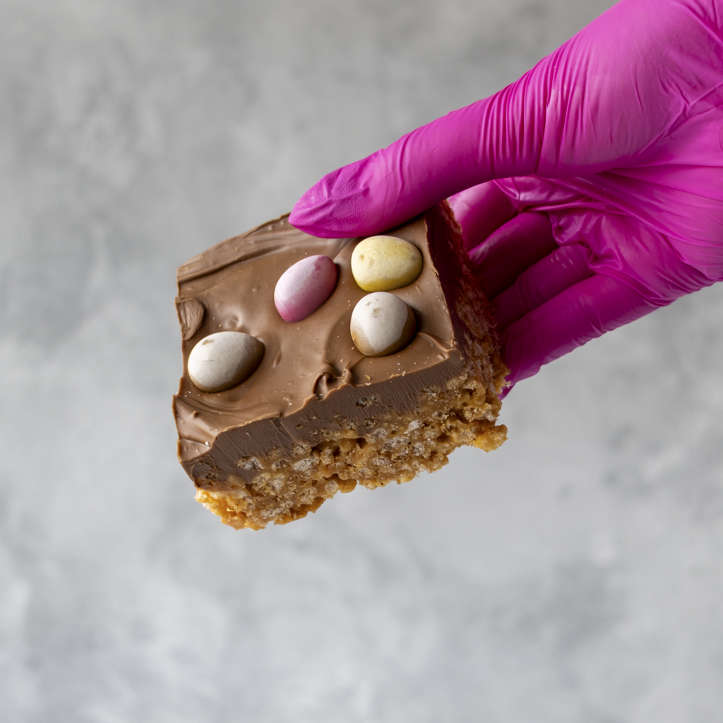 Hand wearing a purple glove holding a Mini Egg Crispie Slab on a gray background