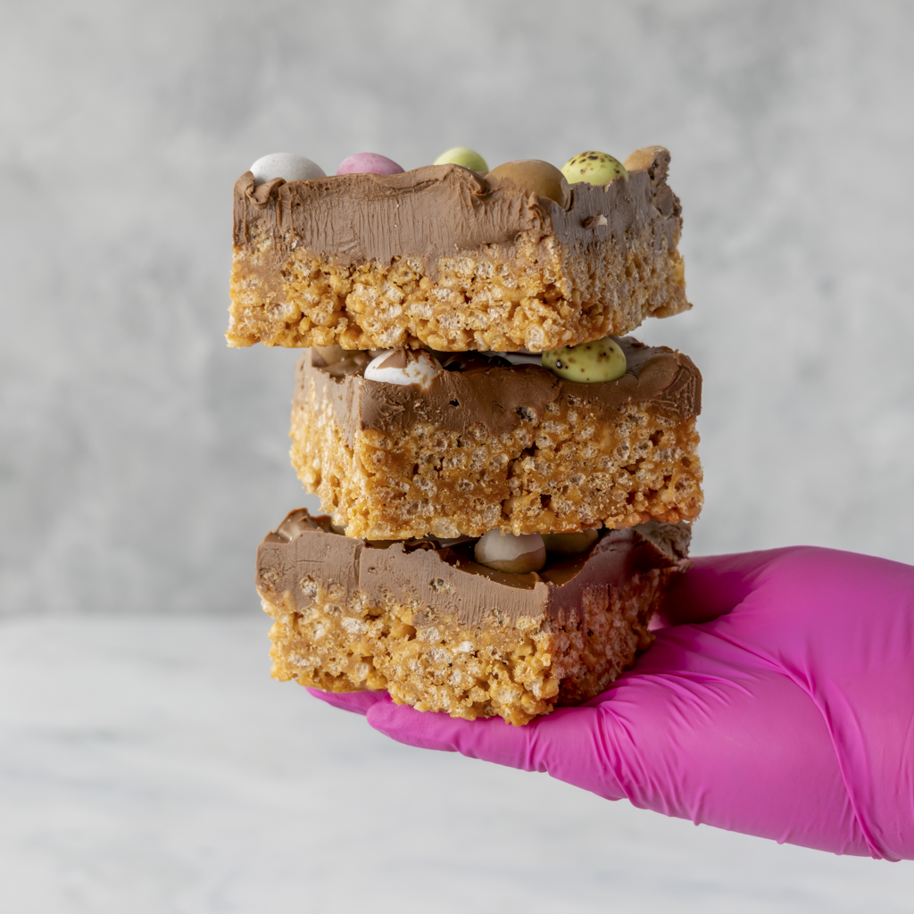 Three layered Mini Egg crispie slab pieces topped with crunchy chocolate mini eggs held in gloved hand
