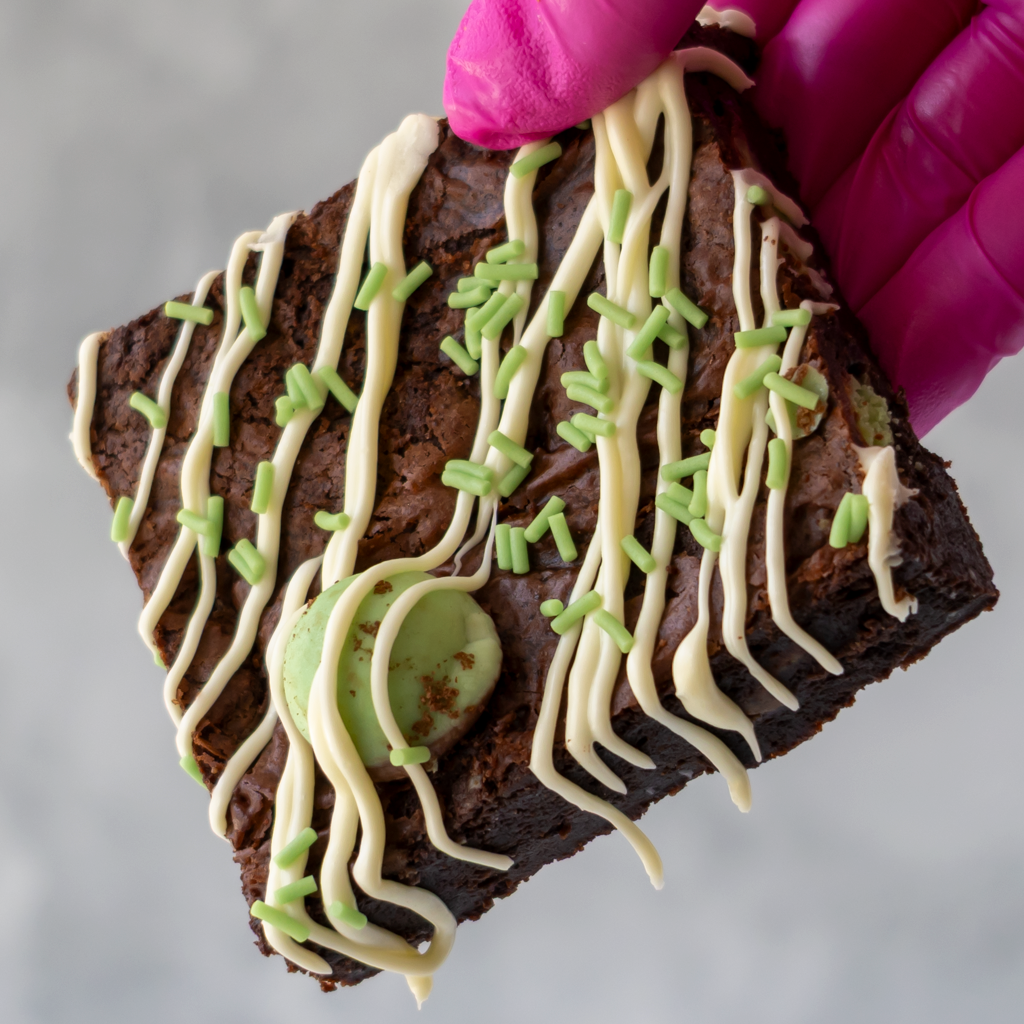 A close-up of a square brownie topped with white chocolate drizzle, green sprinkles, and a mint green cookie piece, held by a hand wearing a bright pink glove against a soft gray background.