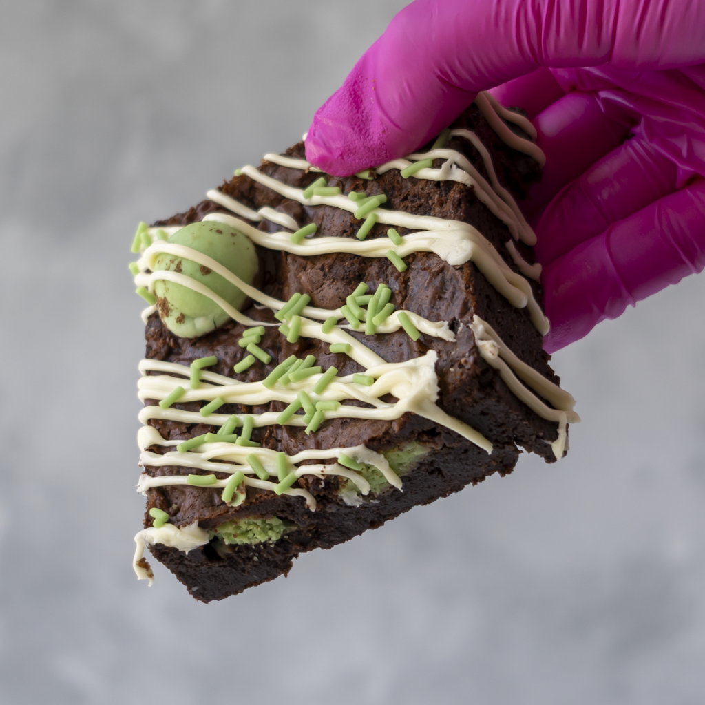 A hand wearing a bright pink glove holds a triangular slice of chocolate brownie decorated with white chocolate drizzle, green sprinkles, and a mint green ball, against a soft gray background.