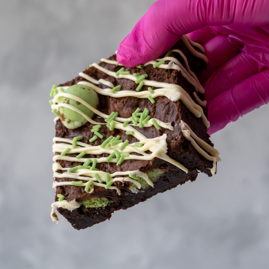 A hand wearing a bright pink glove holds a triangular slice of chocolate brownie decorated with white chocolate drizzle, green sprinkles, and a mint green ball, against a soft gray background.