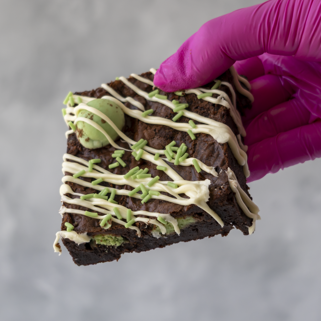 A hand wearing a bright pink glove holds a square chocolate brownie topped with white chocolate drizzle, green sprinkles, and a mint green candy, against a soft gray background.