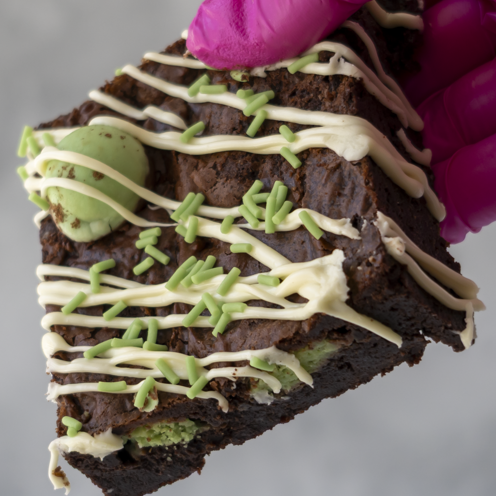 A close-up of a chocolate brownie slice topped with white and cream drizzle, green sprinkles, and a mint green chocolate piece, held by a bright pink gloved hand against a soft gray background.