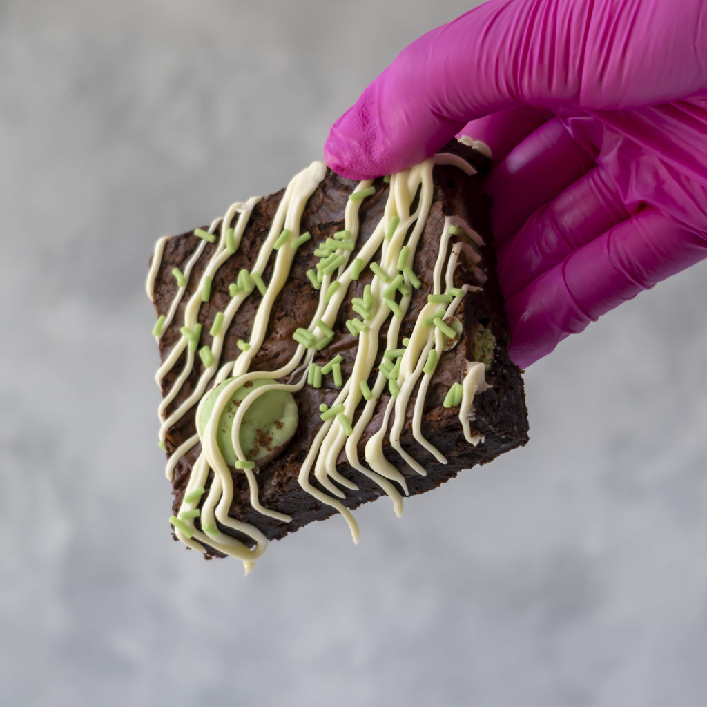 A hand wearing a bright pink glove holds a DoughGirl Monster Mint Brownie Slab topped with white chocolate drizzle, green icing, and a green chocolate ball, against a soft gray background.