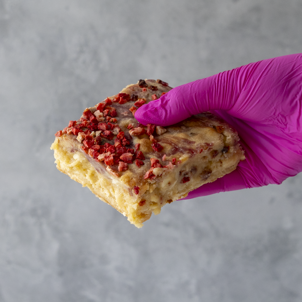 Hand wearing a pink glove holding a Eton Mess Blondie Slab with creamy white chocolate chunks dried red strawberry pieces against a gray background